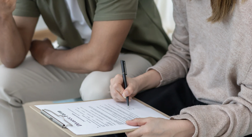 Person filling out a divorce form on a clipboard while another person sits beside them.