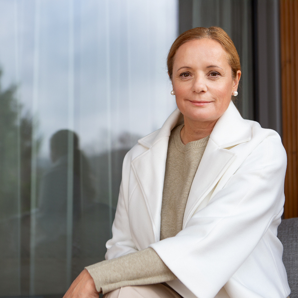 Middle-aged woman with light brown hair wearing a white coat and beige sweater sitting and looking at the camera.
