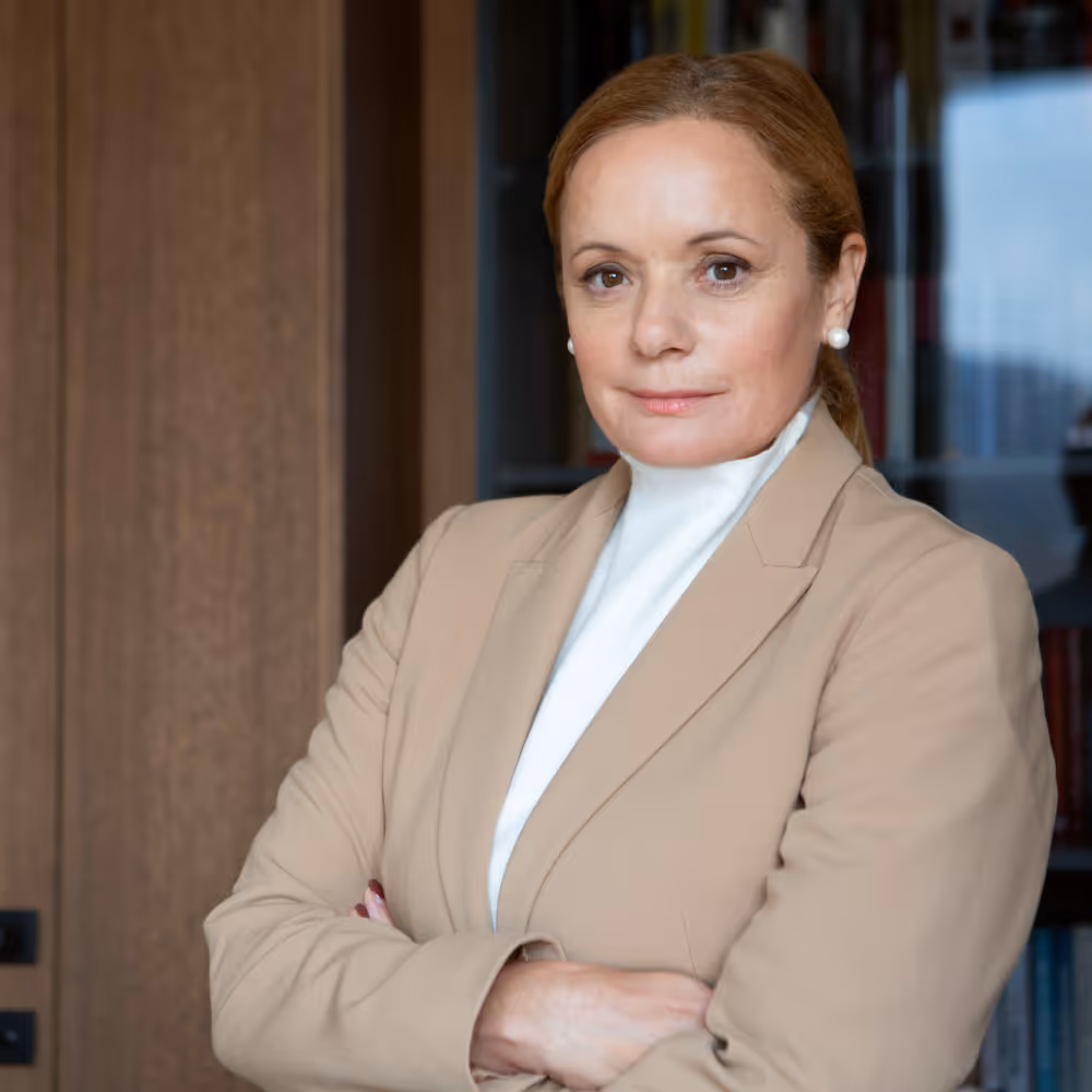 Confident woman in beige blazer with arms crossed standing indoors against wood and glass background.