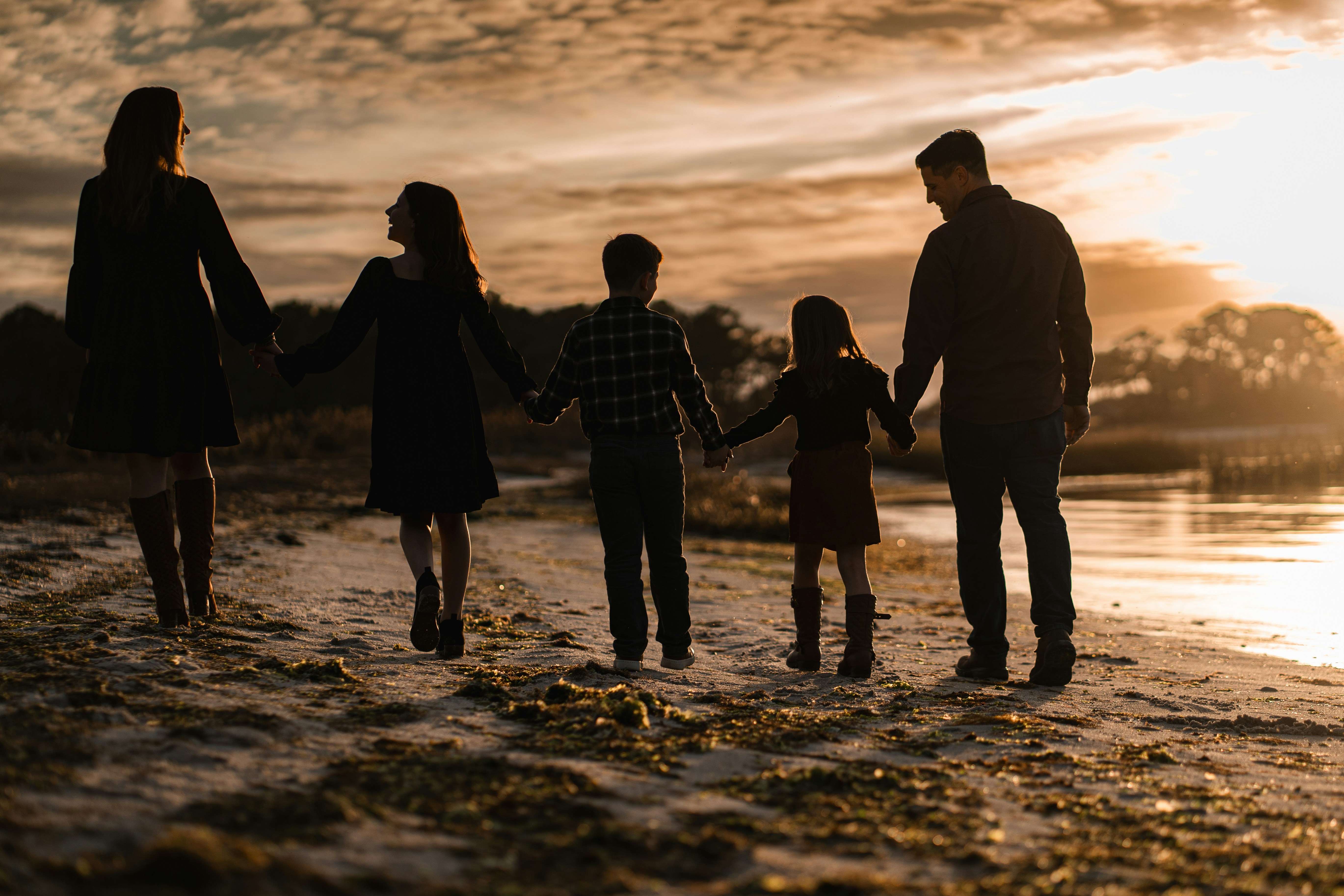 Silhouetted family of five holding hands and walking on a beach at sunset.