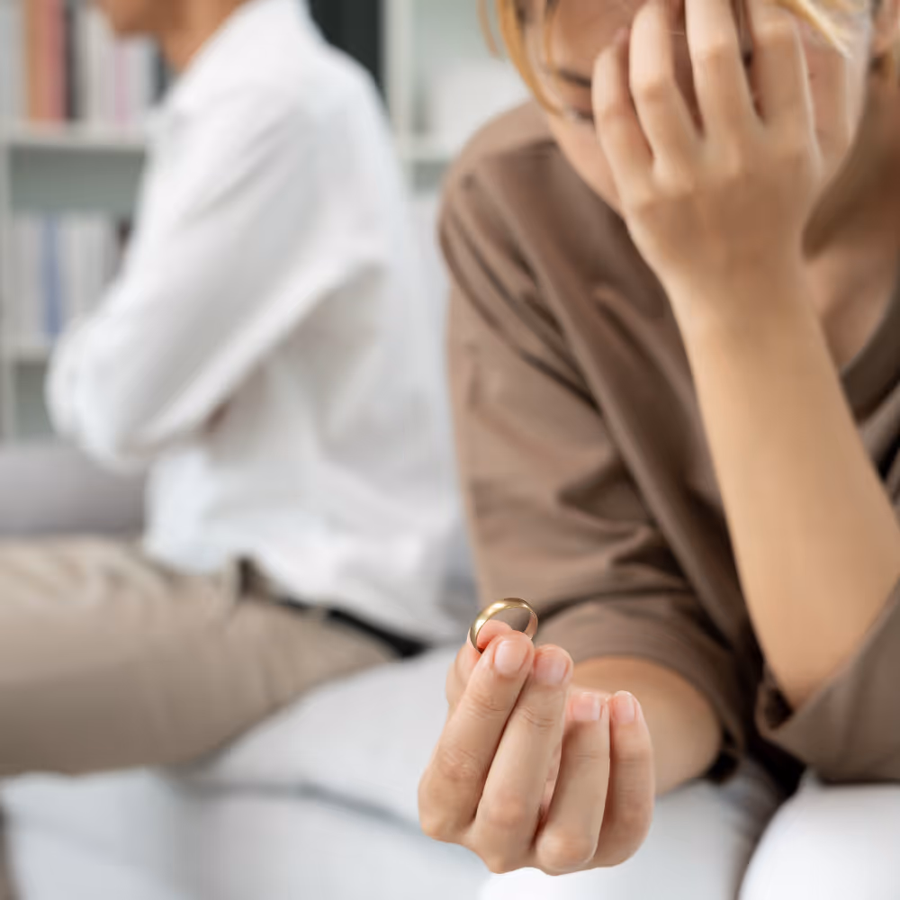 Woman holding a wedding ring looking distressed with a man sitting in the background facing away.