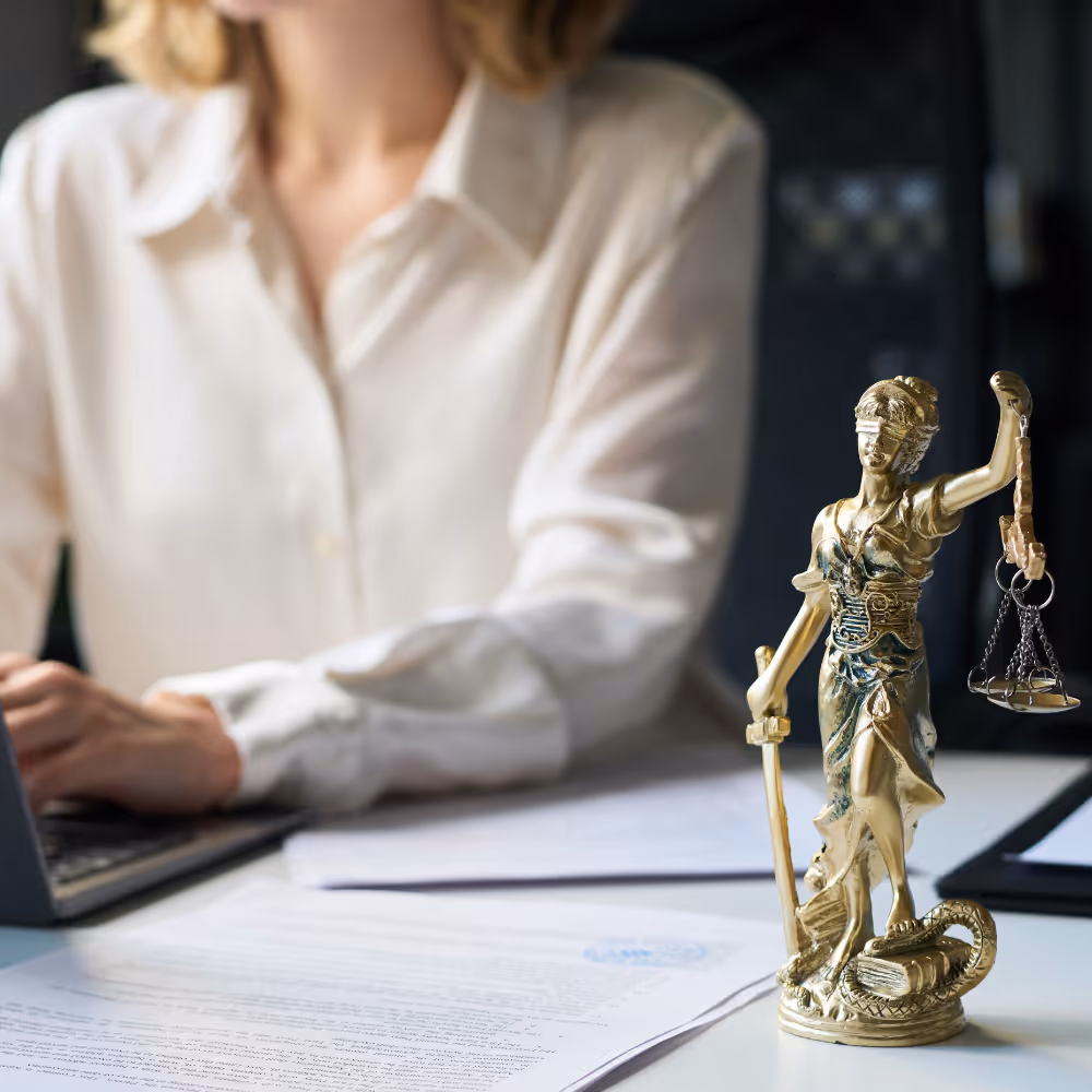 Golden statue of Lady Justice holding scales and a sword on a desk near paperwork and a person typing on a laptop.