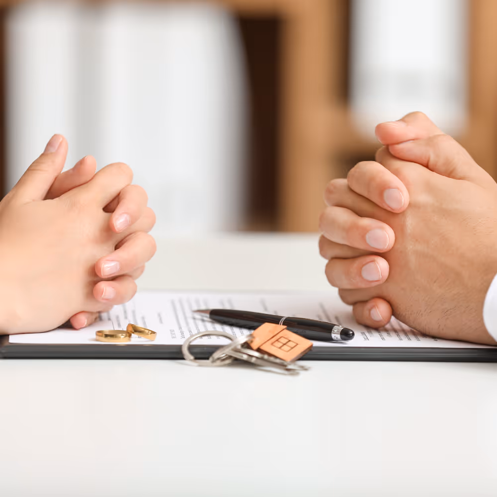 Two people sitting opposite each other with clasped hands over a document, a pen, two wedding rings, and a house-shaped keychain on keys on the table.