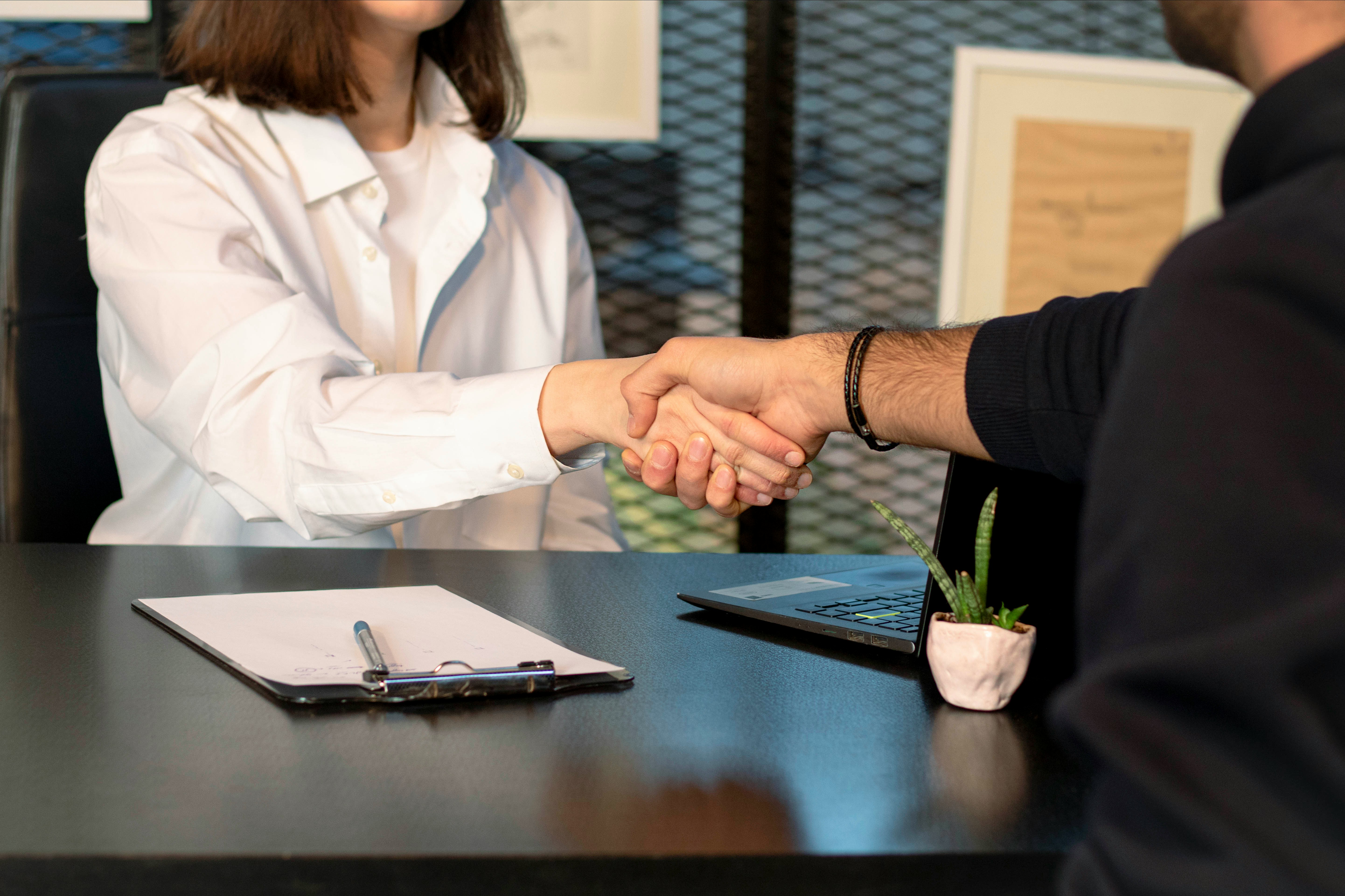 Two people shaking hands across a desk with a clipboard, pen, laptop, and small potted plant.