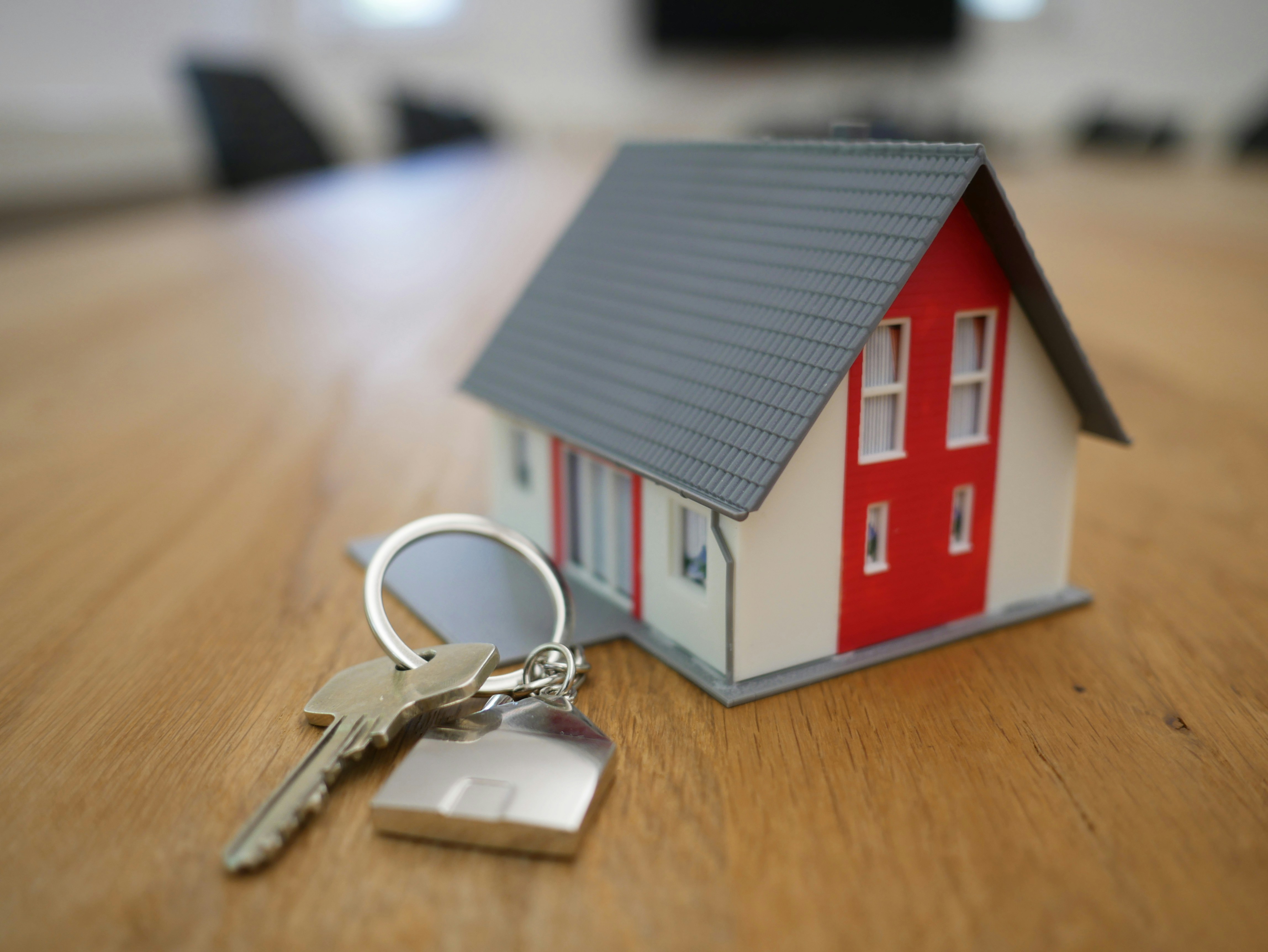 Small model house with red and white walls and gray roof placed on a wooden table next to a key with a house-shaped keychain.